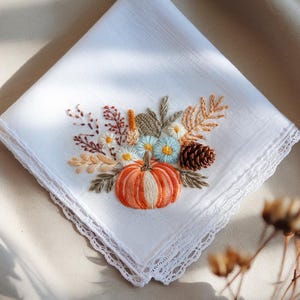 Puede incluir: Pañuelo blanco con diseño bordado de temática otoñal. Presenta una calabaza naranja, una piña y varias flores y follaje en tonos naranja, marrón y azul. Los bordes están acabados con un delicado ribete de encaje.