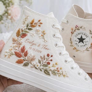 Op de afbeelding: Off-white high-top canvas sneakers met ingewikkeld bloemenborduurwerk in tinten roest, groen en crème. De zijkant toont de geborduurde namen Evelyn & Theo en de datum 10.10.2026. De andere schoen toont een Converse All Star logo-patch.