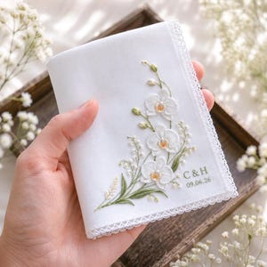 Op de afbeelding: Een witte stoffen zakdoek met delicaat borduurwerk van witte orchideeën en groen blad. De rand is afgewerkt met een witte kanten boord. De tekst C & H en de datum 09.06.26 zijn in groen garen rechtsonder op de zakdoek geborduurd.