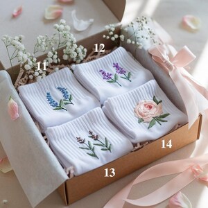 May include: A gift box containing four pairs of white socks, each embroidered with a different floral design in shades of blue, purple, and pink. The box is tied with a pink ribbon and decorated with baby's breath.