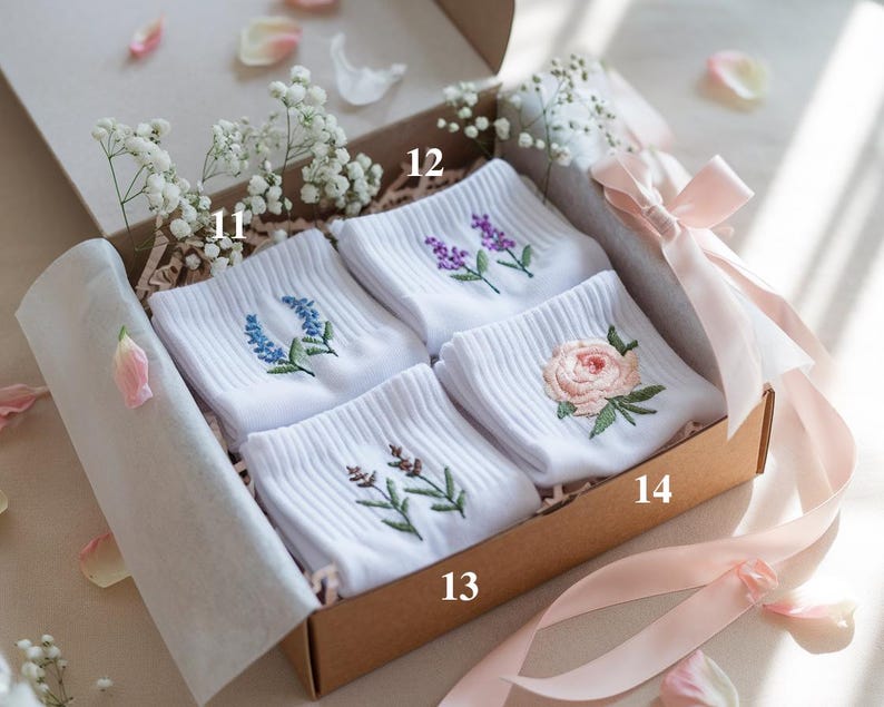 May include: A gift box containing four pairs of white socks, each embroidered with a different floral design in shades of blue, purple, and pink. The box is tied with a pink ribbon and decorated with baby's breath.