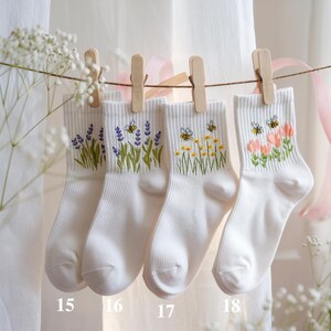 May include: Four pairs of white ankle socks are displayed, each adorned with embroidered floral designs. The socks are suspended by wooden clothespins on a string. The designs include lavender, bees with yellow flowers, and pink flowers.