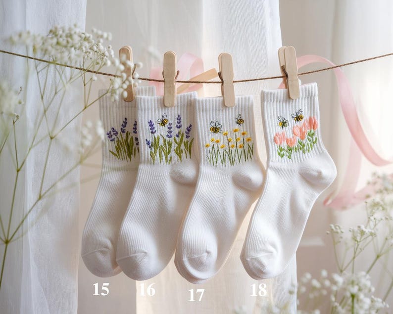 May include: Four pairs of white ankle socks are displayed, each adorned with embroidered floral designs. The socks are suspended by wooden clothespins on a string. The designs include lavender, bees with yellow flowers, and pink flowers.