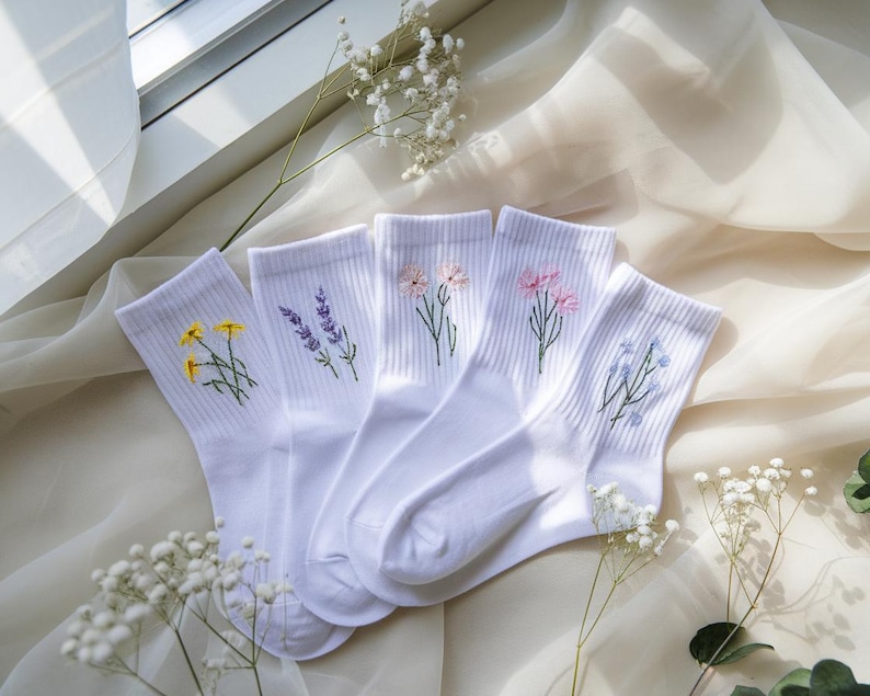 May include: Five pairs of white socks with embroidered floral designs. Each sock features a different flower: yellow, lavender, pink, pink, and blue. The socks are arranged on a cream-colored fabric, with sprigs of white flowers.
