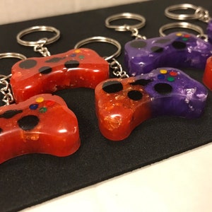 May include: A set of six colorful game controller keychains. The keychains are made of resin and have a glittery finish. Half of the keychains are red with gold glitter and the other half are purple with silver glitter.