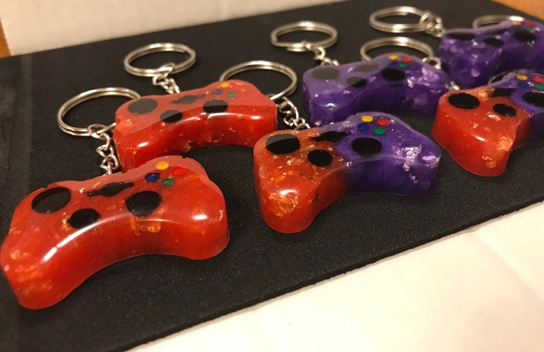 May include: A set of six colorful game controller keychains. The keychains are made of resin and have a glittery finish. Half of the keychains are red with gold glitter and the other half are purple with silver glitter.