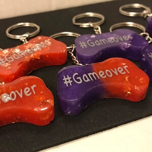 May include: Five purple and red resin game controller keychains with silver key rings. Each keychain has the text "#Gameover" written on it.