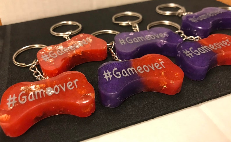 May include: Five purple and red resin game controller keychains with silver key rings. Each keychain has the text "#Gameover" written on it.