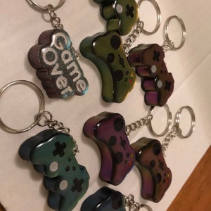 May include: A set of eight iridescent keychains featuring a video game controller design.  The keychains are made of resin and have a holographic finish.  One keychain features the words "Game Over" in white lettering.