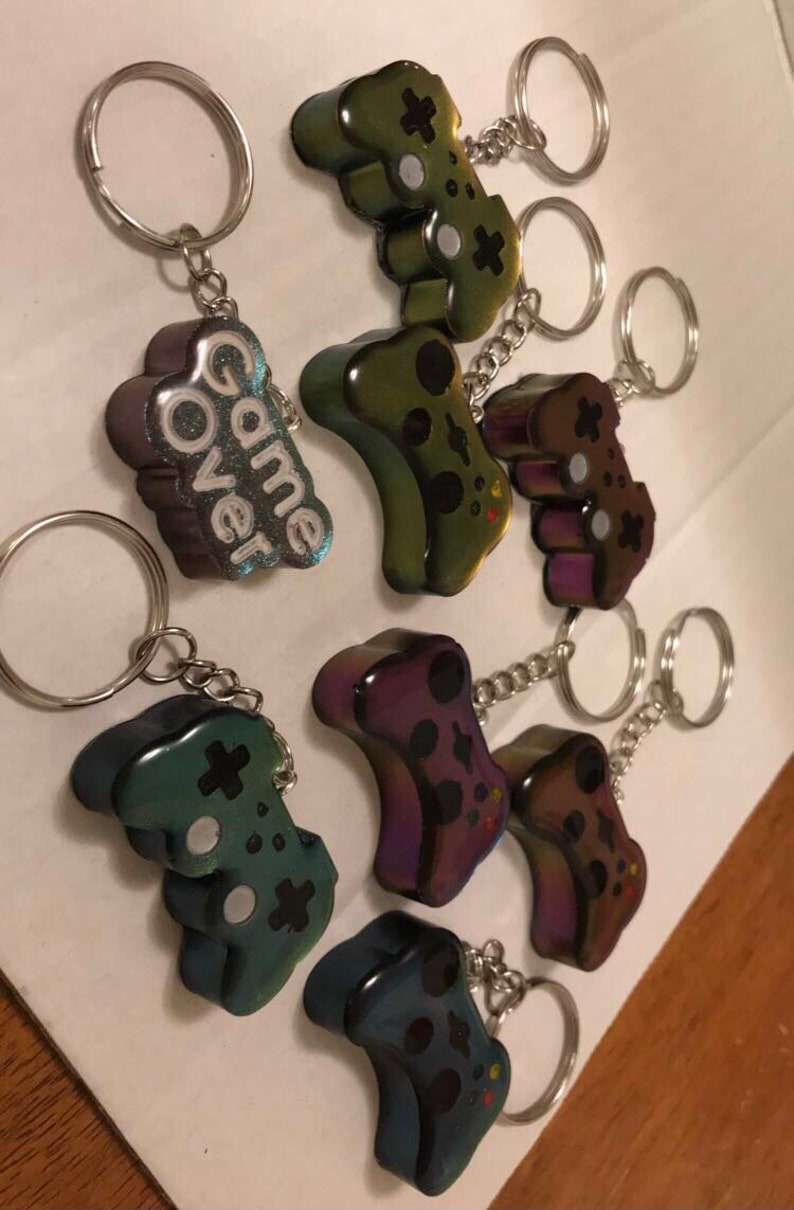 May include: A set of eight iridescent keychains featuring a video game controller design.  The keychains are made of resin and have a holographic finish.  One keychain features the words "Game Over" in white lettering.