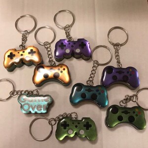 May include: A set of nine keychains featuring different colored game controllers. The controllers are made of resin and have a glossy finish. Some of the controllers have a glittery finish. One keychain features a blue and green controller with the text "Game Over" on it.