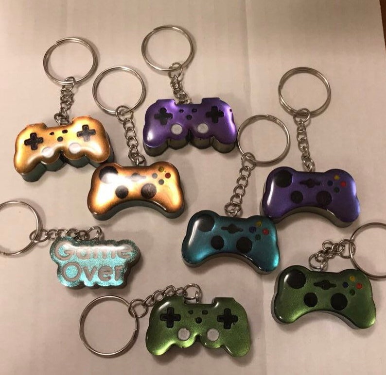 May include: A set of nine keychains featuring different colored game controllers. The controllers are made of resin and have a glossy finish. Some of the controllers have a glittery finish. One keychain features a blue and green controller with the text "Game Over" on it.