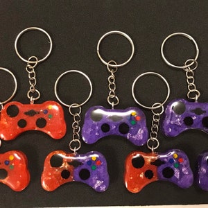 May include: Eight colorful game controller keychains, four red and four purple, with silver key rings and chains. The controllers are made of resin and have a glittery finish.