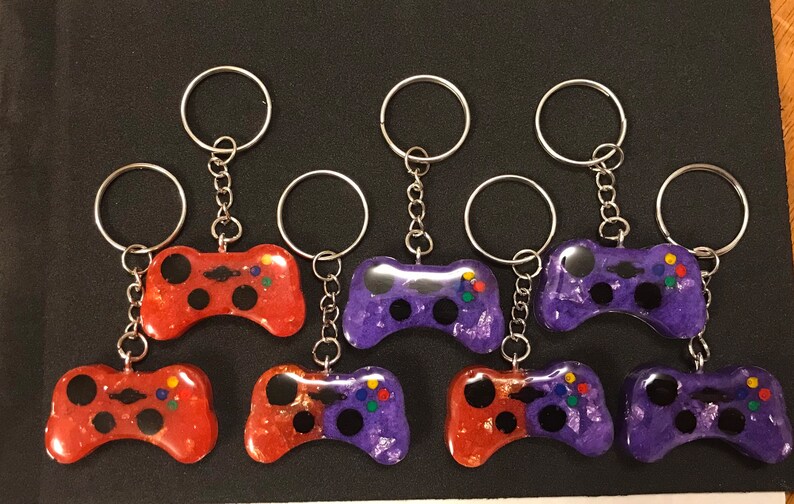 May include: Eight colorful game controller keychains, four red and four purple, with silver key rings and chains. The controllers are made of resin and have a glittery finish.