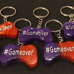May include: Six purple and orange resin game controller keychains with silver glitter and the text "#Gameover" on each.