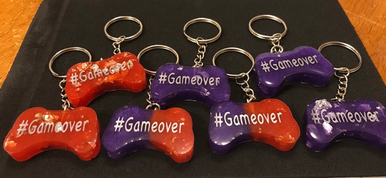 May include: Six purple and orange resin game controller keychains with silver glitter and the text "#Gameover" on each.