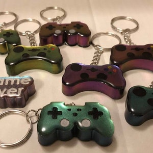 May include: A set of seven iridescent game controller keychains. The keychains are in various colors, including purple, green, and blue. One keychain has the words "Game Over" written on it.