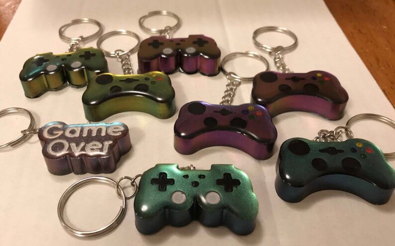 May include: A set of seven iridescent game controller keychains. The keychains are in various colors, including purple, green, and blue. One keychain has the words "Game Over" written on it.