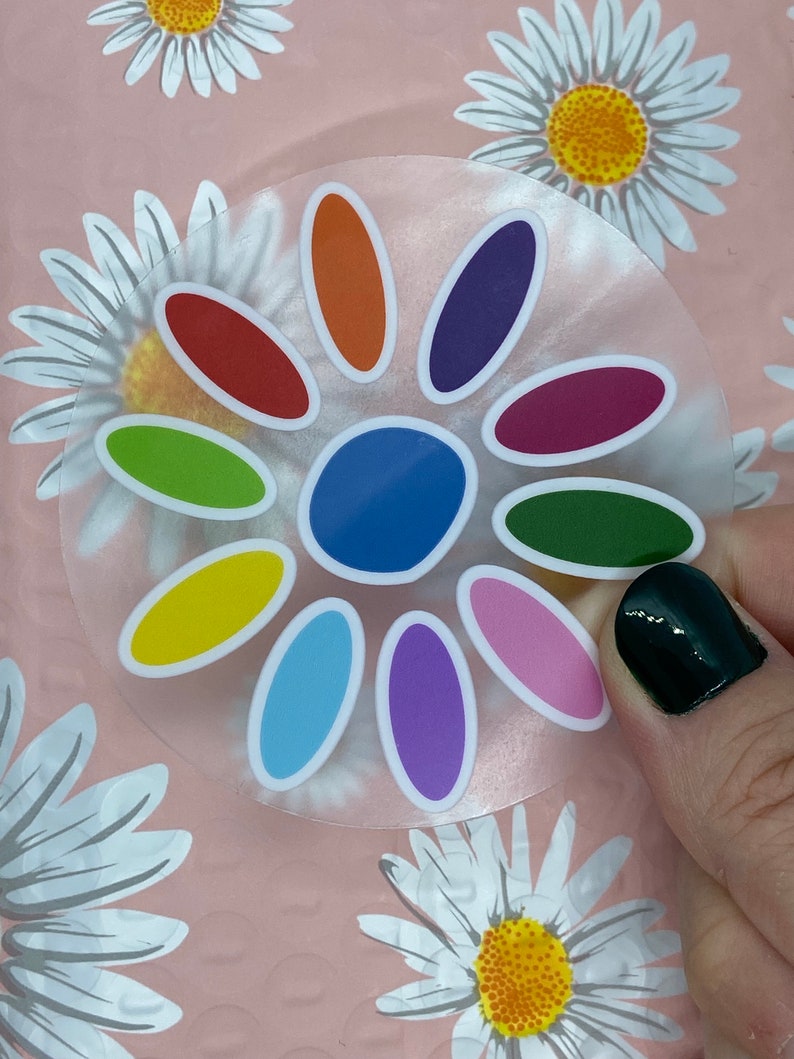 May include: A clear sticker with a colorful flower design. The flower has 12 petals in various colors, including red, orange, yellow, green, blue, purple, and pink. The center of the flower is blue.