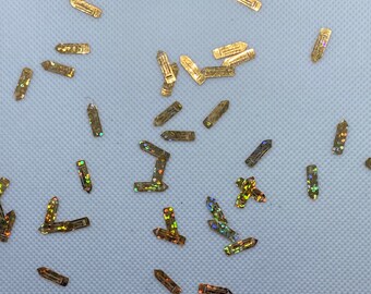 May include: A scattering of gold pencil-shaped glitter on a white background. The glitter has a holographic finish, creating a rainbow effect.