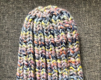 May include: A colorful knitted beanie hat with a cable knit pattern. The hat is made with a blend of blue, pink, yellow, and white yarn.