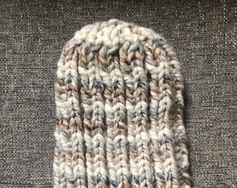 May include: A white, brown, and gray knitted beanie hat. The hat is made with a chunky yarn and has a ribbed texture.