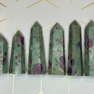 May include: Seven green and pink ruby in zoisite crystal points, also known as ruby in fuchsite, arranged in a row on a white background with a gold geometric pattern.