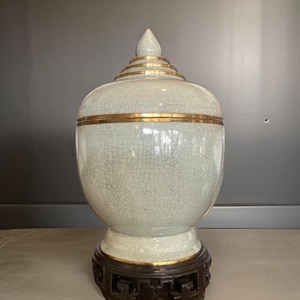 Könnte beinhalten: Eine weiße Keramikvase mit goldener Verzierung und einem spitzen Deckel. Die Vase steht auf einem Holzsockel mit aufwendigen Schnitzereien.
