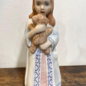 Könnte beinhalten: Porzellanfigur eines jungen Mädchens in einem weißen Gewand, das einen kleinen Teddybären hält. Das Gewand hat blaue Verzierungen mit einem sich wiederholenden Muster. Das Mädchen hat rotes Haar und steht auf einer Holzoberfläche.