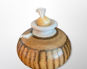 May include: A small, round, ceramic oil lamp with a brown and white glaze. The lamp has a white wick holder and a wooden wick.