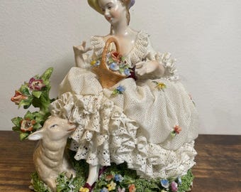 May include: A porcelain figurine of a woman in a white dress with lace and ruffles, holding a basket of flowers. A lamb is beside her on a grassy base with colorful flowers. The woman wears a yellow hat and has a purple flower in her hair.
