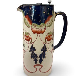 Puede incluir: Jarra alta de color crema con la parte superior y el asa de color azul oscuro. La jarra presenta un diseño floral con flores rojas y verdes y hojas azules. Se ven detalles dorados en el borde y el asa. La jarra tiene una tapa plateada con un botón blanco.