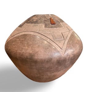 May include: A handcrafted, brown ceramic vessel with a rounded shape and a flat top. The top features a geometric design and a small, triangular wooden accent. The vessel's surface has a textured appearance, showcasing the natural variations of the clay.