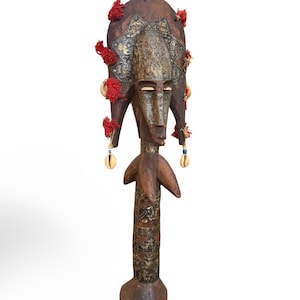 Puede incluir: Una máscara de madera tallada con un tocado alto y redondeado adornado con borlas rojas y detalles metálicos. La máscara presenta un rostro sereno con una nariz larga y ojos cerrados. Conchas cuelgan de los lados. La máscara está montada sobre un soporte de madera.