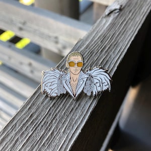 May include: A white enamel pin featuring a cartoon portrait of a man wearing a white sequined jacket with feathered shoulders and yellow sunglasses.