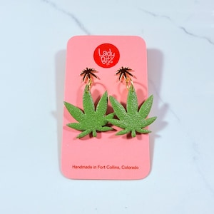 May include: Green marijuana leaf earrings with gold hoops and a gold star charm. The earrings are on a pink card with the text "Lady Bits Co." and "Handmade in Fort Collins, Colorado."
