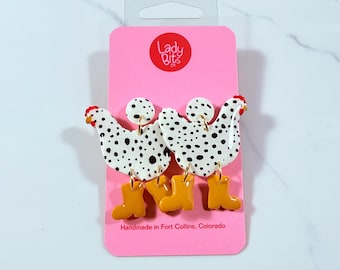 May include: A pair of white and black spotted chicken earrings with yellow boots. The earrings are handmade in Fort Collins, Colorado by Lady Bits Co.