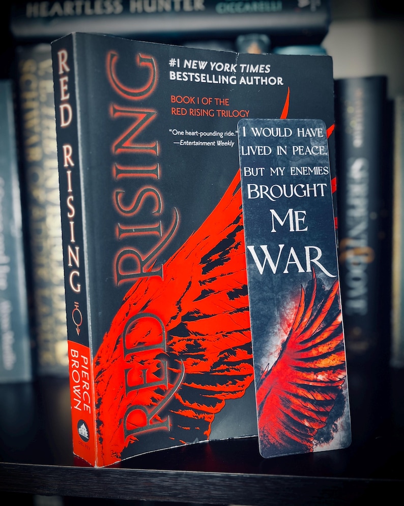 May include: A hardcover book titled "Red Rising" by Pierce Brown, with a red and black cover design. A bookmark with the quote "I would have lived in peace, but my enemies brought me war" is next to the book. The book is part of the Red Rising trilogy.