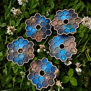 May include: Five hexagonal wooden coasters with a galaxy design. Each coaster features a blue and brown color scheme with white star details. The coasters are arranged on a bed of green leaves and white flowers.