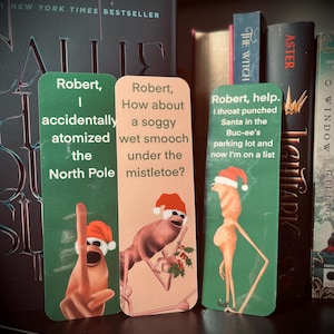 Puede incluir: Tres marcapáginas humorísticos con temática navideña con personajes de dibujos animados que llevan gorros de Papá Noel. Los marcapáginas son verdes y rosas con texto que dice "Robert, accidentalmente atomizé el Polo Norte", "Robert, ¿Qué tal un beso empapado bajo el muérdago?" y "Robert, ayuda. Le di un puñetazo a Papá Noel en el estacionamiento de Buc-ee y ahora estoy en una lista."