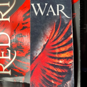 May include: A bookmark featuring a red and black wing design with the words "ME WAR" in white. The bookmark is rectangular and has a dark background, with the wing design taking up most of the space.