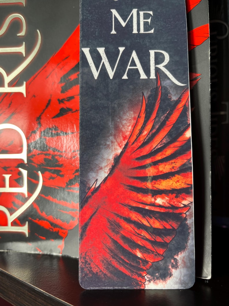 May include: A bookmark featuring a red and black wing design with the words "ME WAR" in white. The bookmark is rectangular and has a dark background, with the wing design taking up most of the space.