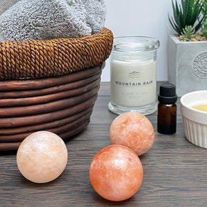 May include: Three orange Himalayan salt balls, a clear glass candle with a white label that reads "Mountain Rain", a small brown glass bottle, and a white ceramic bowl on a wooden surface.
