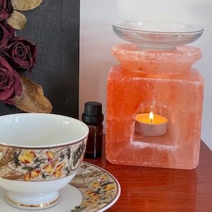 May include: A pink Himalayan salt lamp with a glass bowl on top and a small tea light candle in the centre. The lamp is on a wooden surface with a teacup and saucer next to it.