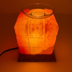 May include: A glowing orange Himalayan salt lamp with a clear glass dish on top. The lamp has a rectangular shape with a grid-like texture and sits on a dark wooden base. A black power cord extends from the base.