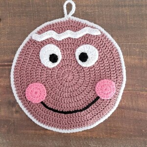 May include: A crocheted gingerbread man hot pad with a pink face, white icing, and black eyes. The hot pad has a loop for hanging.