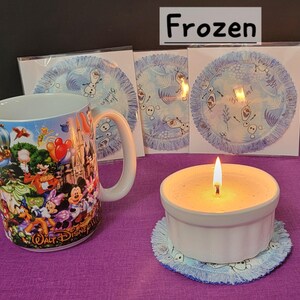 Puede incluir: Una taza de cer&aacute;mica blanca con un dise&ntilde;o tem&aacute;tico de Frozen que presenta personajes de la pel&iacute;cula. La taza est&aacute; sobre una superficie morada junto a un portavelas de cer&aacute;mica blanca con una vela encendida dentro. Hay tres posavasos azules y blancos con un dise&ntilde;o de Frozen en el fondo.