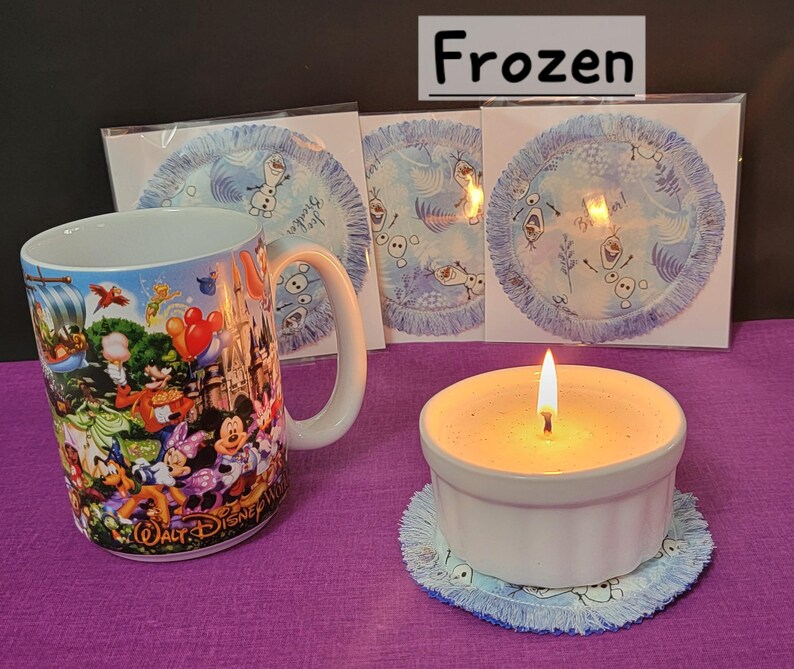 Puede incluir: Una taza de cer&aacute;mica blanca con un dise&ntilde;o tem&aacute;tico de Frozen que presenta personajes de la pel&iacute;cula. La taza est&aacute; sobre una superficie morada junto a un portavelas de cer&aacute;mica blanca con una vela encendida dentro. Hay tres posavasos azules y blancos con un dise&ntilde;o de Frozen en el fondo.