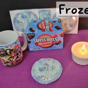 Puede incluir: Un set de regalo de Frozen con una taza con personajes de la pel&iacute;cula, una caja de mezcla de chocolate caliente Swiss Miss, una vela y un posavasos de tela azul y blanco con un dise&ntilde;o de copo de nieve. El posavasos tiene el texto "Let It Snow".