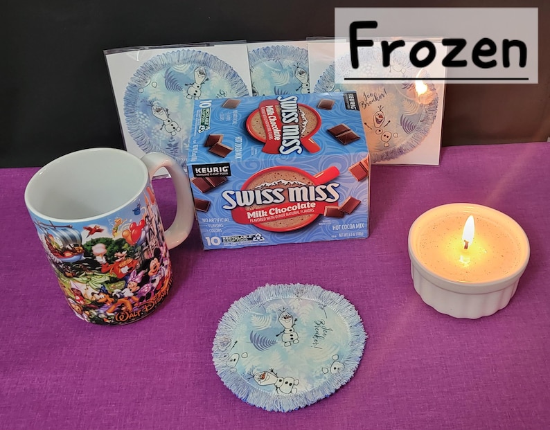 Puede incluir: Un set de regalo de Frozen con una taza con personajes de la pel&iacute;cula, una caja de mezcla de chocolate caliente Swiss Miss, una vela y un posavasos de tela azul y blanco con un dise&ntilde;o de copo de nieve. El posavasos tiene el texto "Let It Snow".